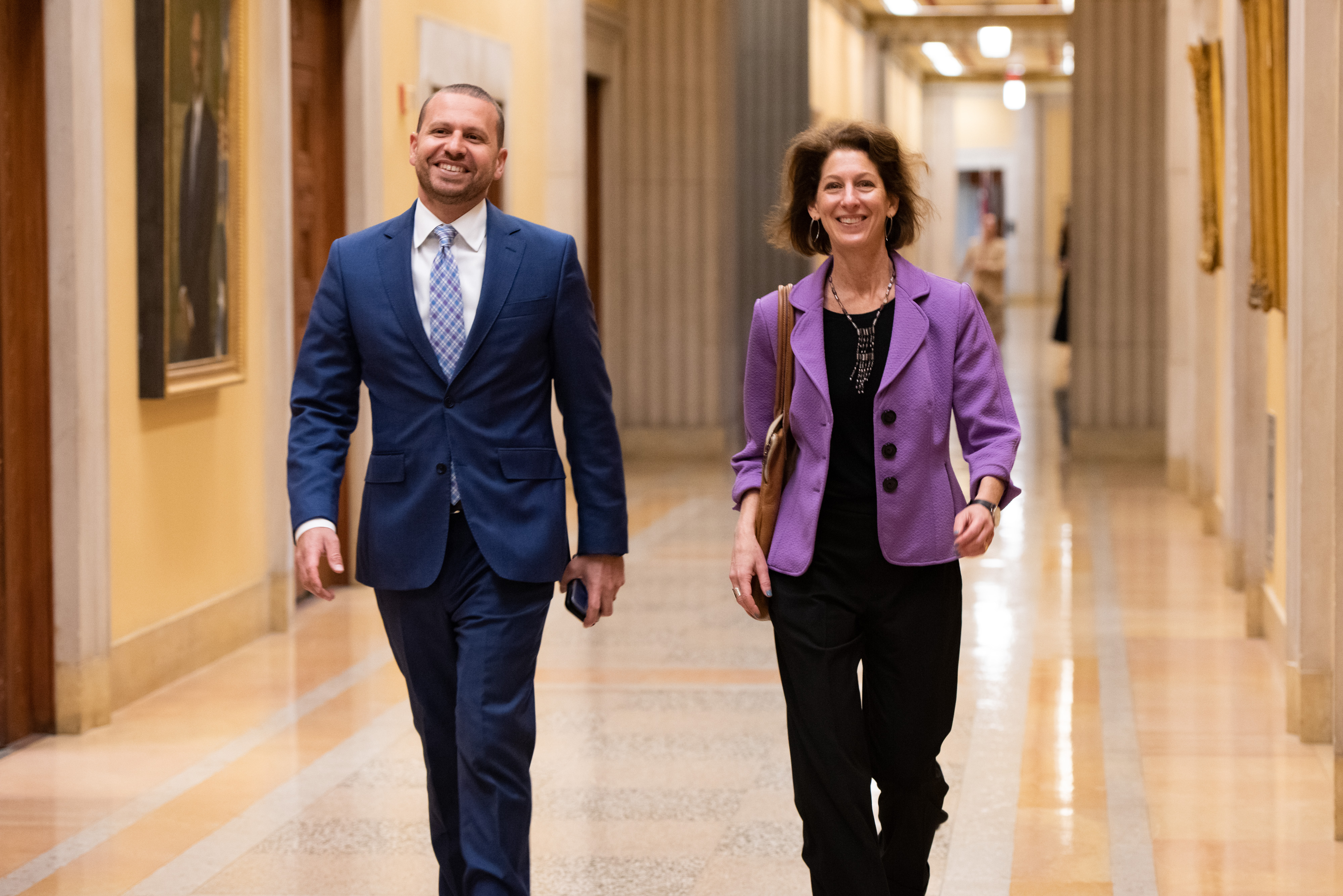 Assistant Attorney General Amy L. Solomon walking alongside Principal Deputy Assistant Attorney General Brent Cohen in a government building hallway, both smiling and dressed in business attire.