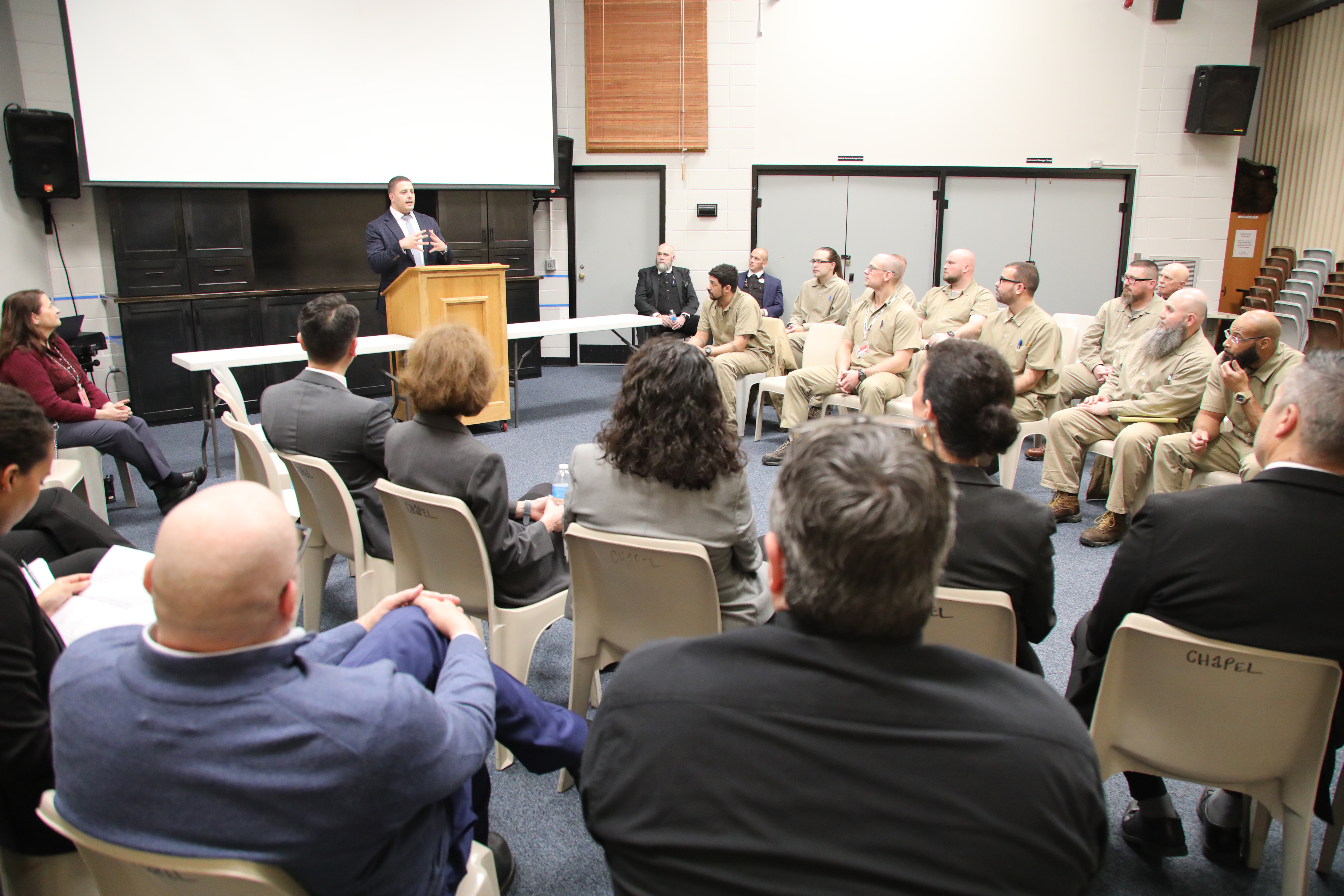 Speaker Addressing Group at Correctional Education Event A speaker stands at a podium addressing a mixed group of inmates and officials seated in a large room during a correctional education event. The audience includes both uniformed inmates and formally dressed officials, attentively listening to the presentation.