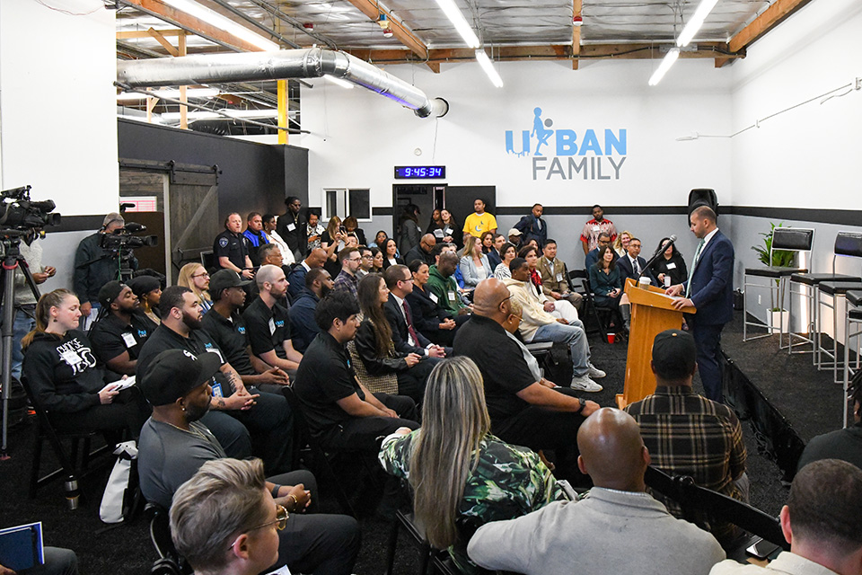 A crowded room with diverse attendees listening to a speaker at a podium. The Urban Family logo is visible on the wall, and various equipment including cameras and a digital clock can be seen.