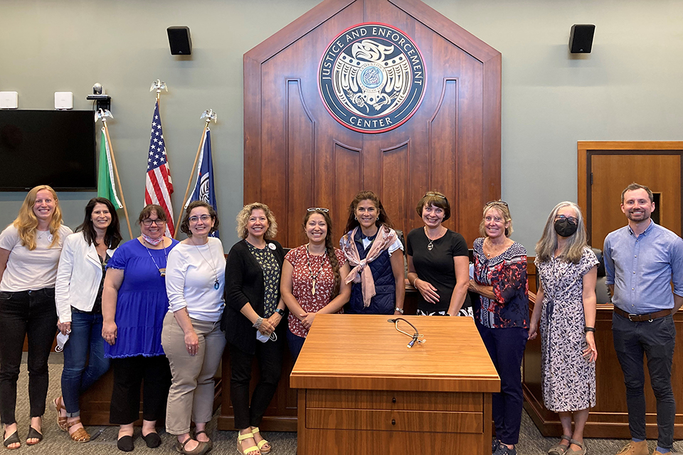 Center for Children & Youth Justice CCYJ Group A group of eleven people standing in a room with a wooden podium and a large emblem on the wall behind them that reads 'Justice and Enforcement Center.' The group is positioned in front of three flags: one green, one American, and one blue. The room has a formal setting with a wooden door and a television screen on the left side.