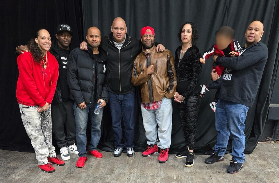 The Black Rose Collective Seven people standing together in front of a black curtain. They're dressed in various outfits, including a red hoodie, a black puffer jacket, a brown leather jacket, and a black patterned outfit. One person is holding a child dressed in red and black. The group appears to be posing for a casual photo, with some wearing sneakers and others wearing casual shoes. The floor is wooden.