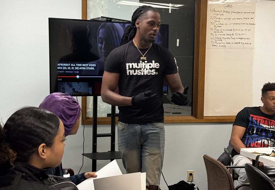 Pro Se Potential A man wearing a 'multiple hustles' t-shirt speaks to a small audience in a classroom setting. A woman in the foreground holds papers, while a TV screen and whiteboard are visible in the background, suggesting an educational or training environment.