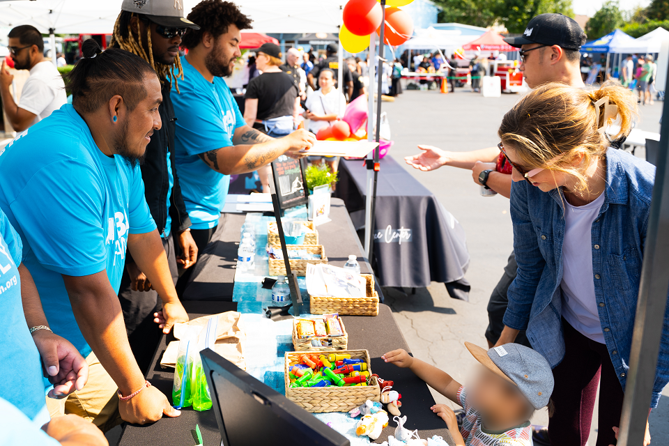 Urban Family Volunteers in bright blue shirts engage with attendees at a lively street fair booth. The table displays colorful items and informational materials, while balloons and tents are visible in the background, creating a festive atmosphere.