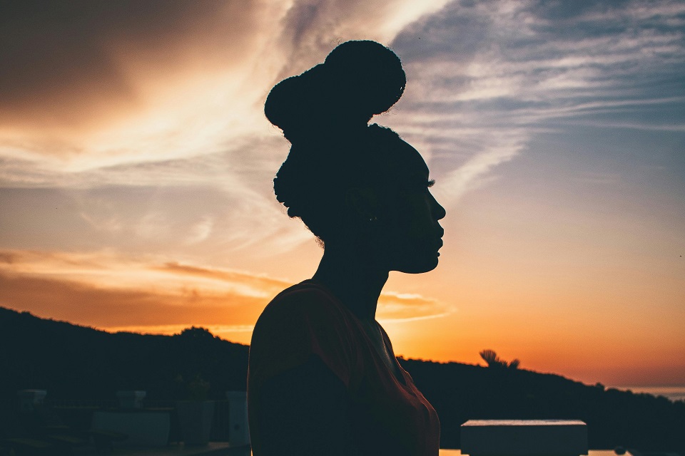 A silhouette profile of a person with their hair in a top bun, photographed against a dramatic orange and purple sunset sky with wispy clouds. Hills and water can be seen in the background.