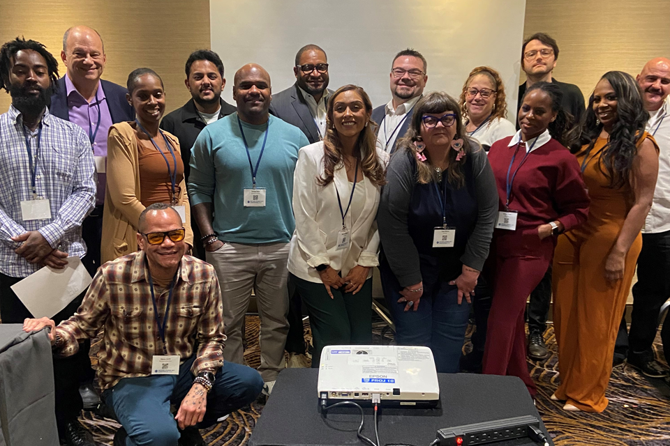 National Reentry Housing Incubator Grantees with Impact Justice Staff at Second Chance Act Conference A group photo of approximately fourteen people from the National Reentry Housing Incubator program. The group is arranged in front of a neutral wall, with a projector visible on a table in the foreground. Attendees are dressed in professional attire ranging from business casual to formal, including outfits in colors like purple, turquoise, white, burgundy, and orange.