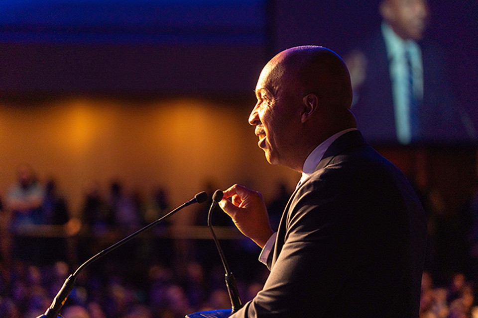 A man in a dark suit speaking at a podium, photographed in profile against a dark background with warm lighting.