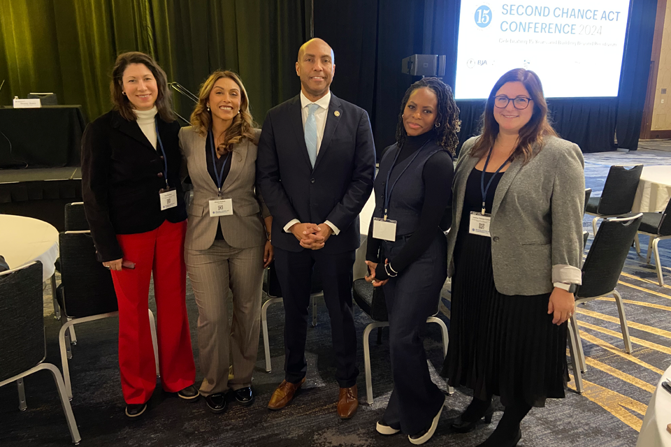 Impact Justice and BJA Staff at Second Chance Act Conference Five staff members from Impact Justice and the Bureau of Justice Assistance pose in front of a Second Chance Act Conference banner. They are wearing professional attire including blazers, suits, and professional dresses in colors including black, red, gray, and navy.