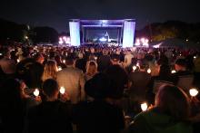 Night scene with crowd holding lit candles facing lighted stage.